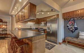 A modern kitchen with wooden cabinets, a large island with stools, a stainless steel refrigerator, and a microwave.