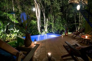 Un espace extérieur avec une piscine, des chaises longues et une petite table, le tout entouré d’une végétation luxuriante et d’arbres.