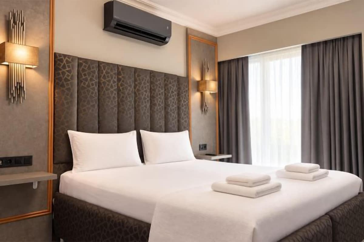 A hotel room with a large bed, two bedside tables, an air conditioning unit, and a window with curtains.