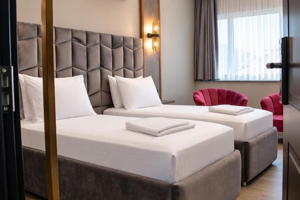 A hotel room with two beds, a grey upholstered headboard, white bedding, a red armchair, and a window with curtains.