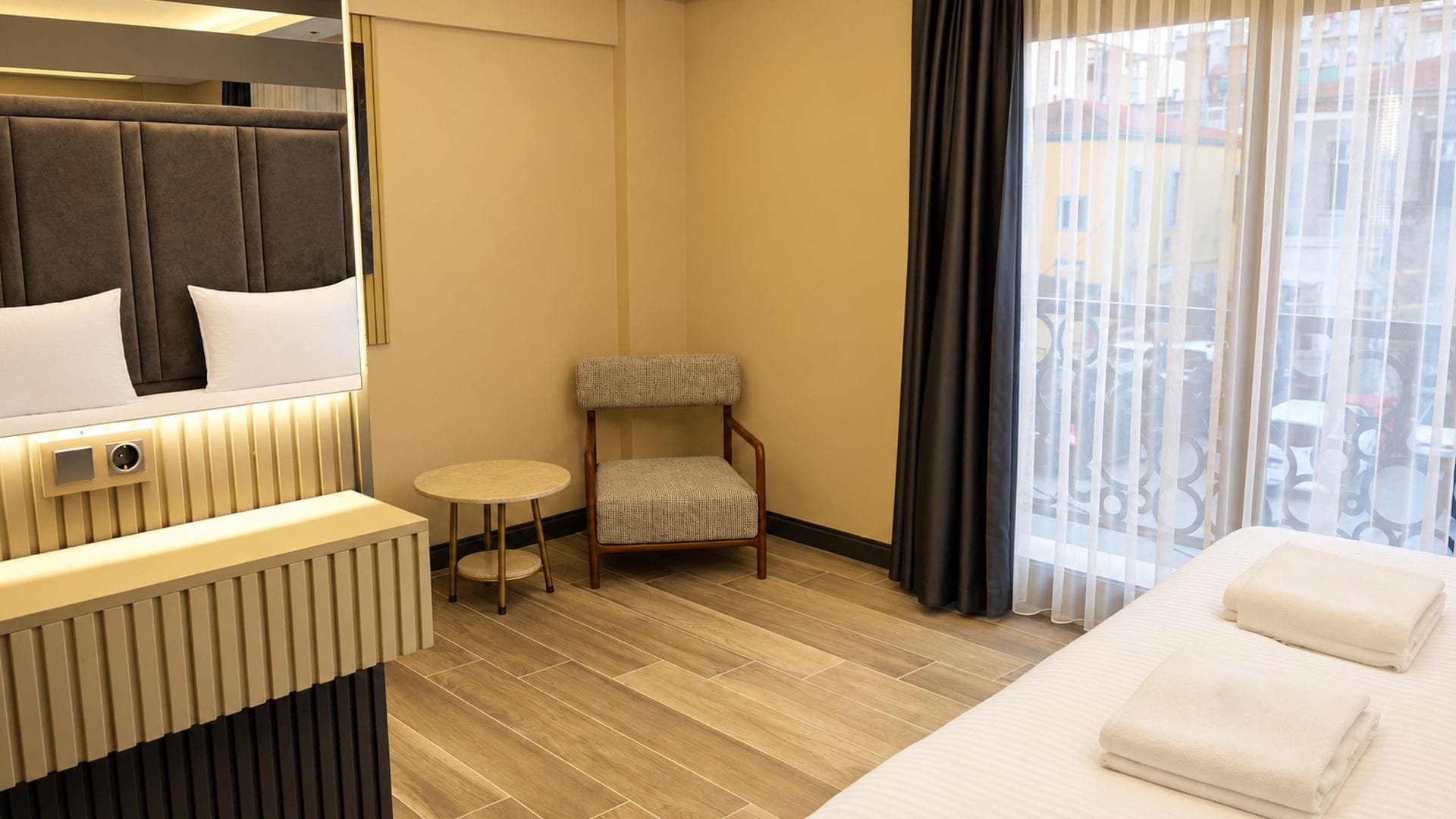A hotel room with a bed, a chair, a small table, a mirror, and a window with curtains.