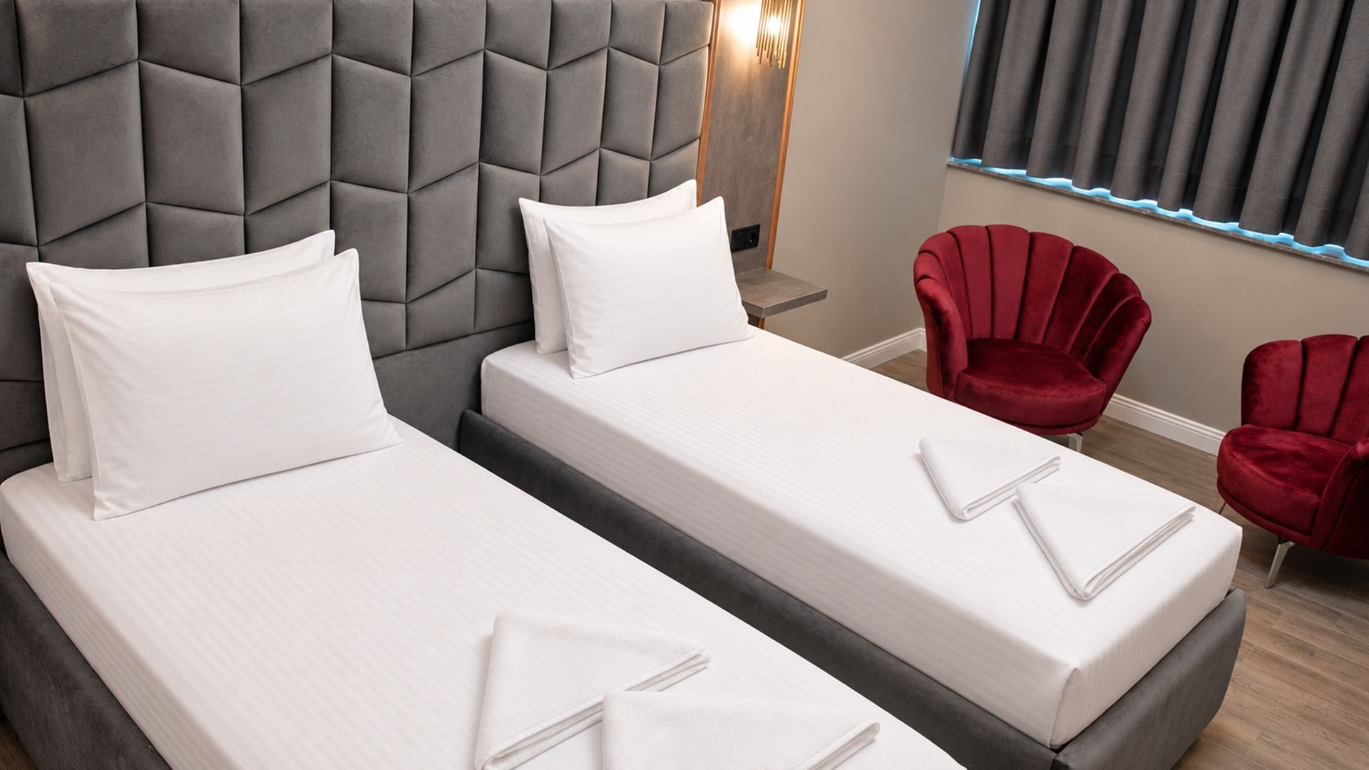 A hotel room with two beds, a grey headboard, red chairs, and a window with curtains.