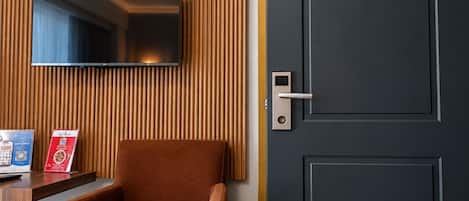 Ein Hotelzimmer mit an einer Holzvertäfelung angebrachtem Fernseher, einer Tür mit moderner Verriegelung, einem braunen Ledersessel und einem kleinen Tisch mit einer Broschüre.