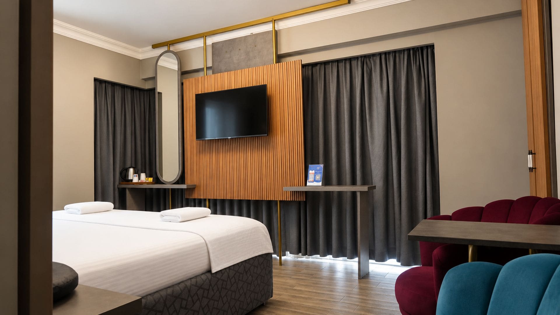 A hotel room with a bed, a TV mounted on a wooden panel, a small table, and a red chair.