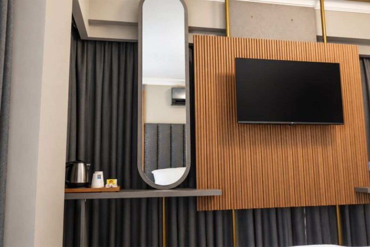 Ein modernes Hotelzimmer mit einem an einer Holzplatte montierten Flachbildfernseher, einem großen Spiegel links und einem kleinen Tisch mit Wasserkocher und Tasse.