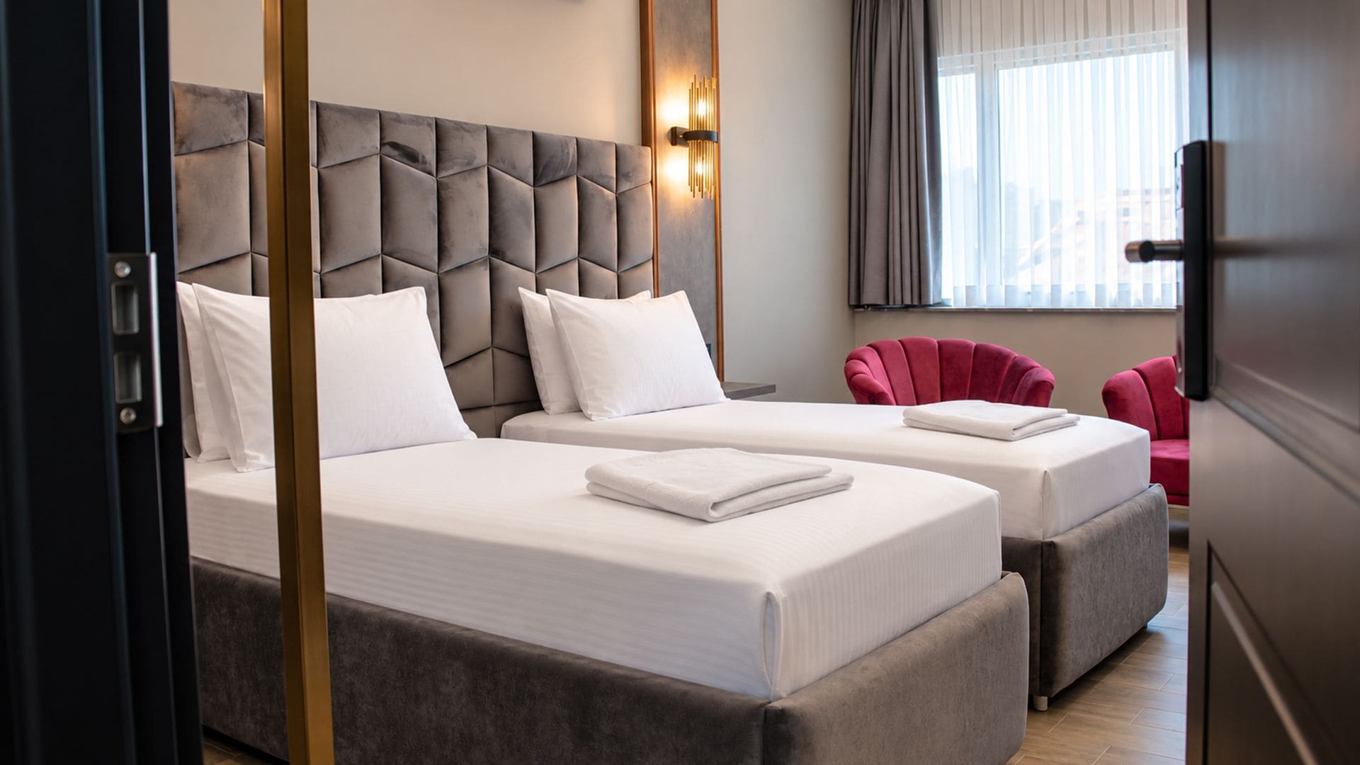 Habitación de hotel con dos camas, un cabecero tapizado en gris, un sillón rojo y una ventana con persianas.