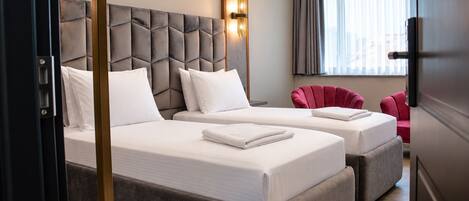 Habitación de hotel con dos camas, un cabecero tapizado en gris, un sillón rojo y una ventana con persianas.