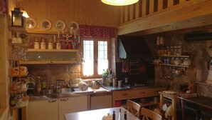 Maalaishenkinen keittiö, jossa on puuseinät, kivitaso ja erilaisia keittiövälineitä. Siellä on hyllyjä, joilla on purkkeja ja lautasia, sekä pesuallas ja ikkuna, jossa on verhot.