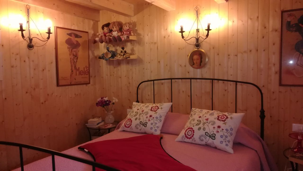 Une pièce aux murs lambrissés, avec un lit, des coussins à motifs floraux, une couverture rouge, une table de chevet ornée d’un vase de fleurs et des étagères murales décoratives.