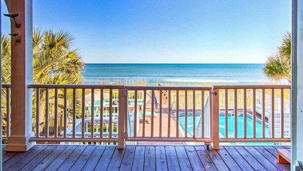 A wooden deck with a view of a beach and pool area.