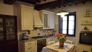 A kitchen with white cabinets, a stove, a sink, and a dining table with chairs. There is a window with curtains and a vase with flowers on the table. - La Petronilla Montepetriolo - Apartment Delight (Monte Petriolo)