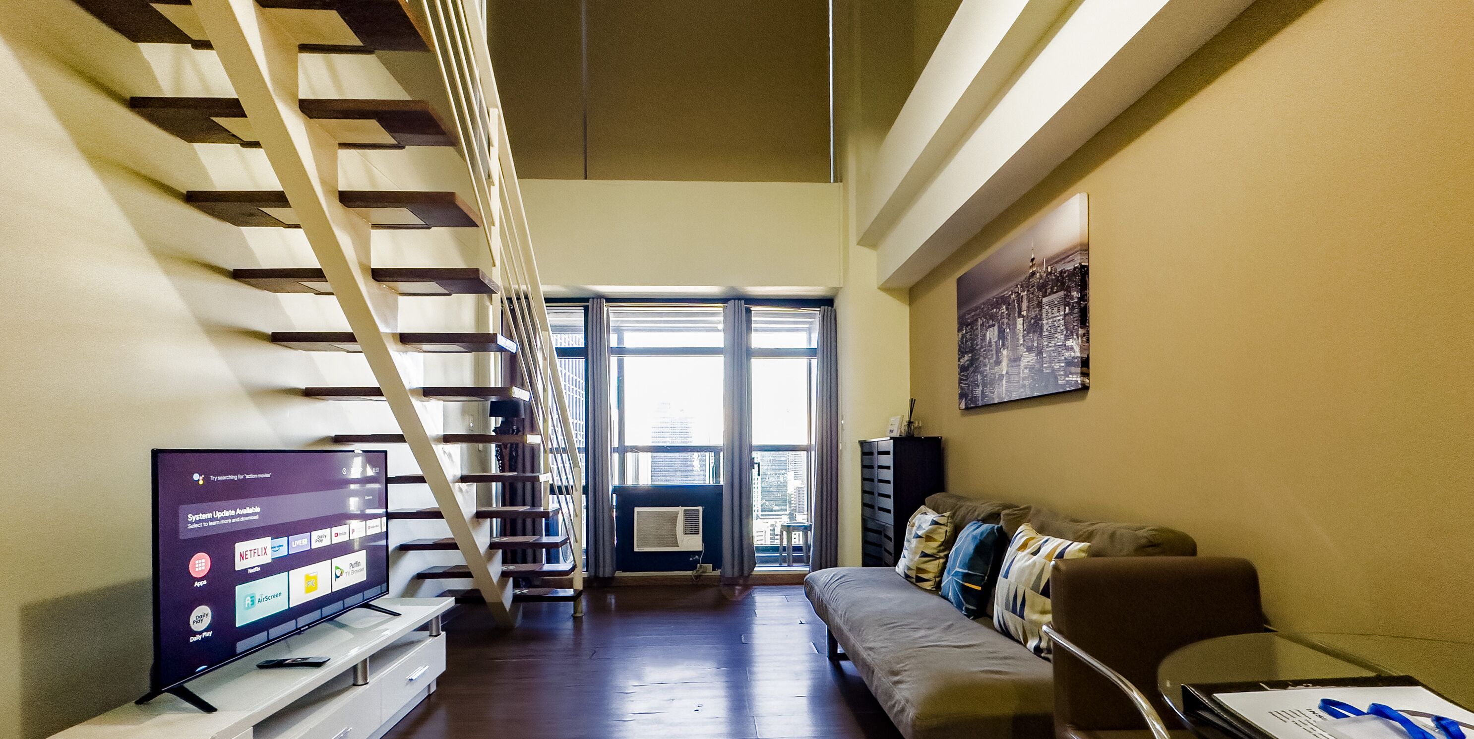 A modern living room with a staircase, a flat-screen TV, a sofa, and a city view.