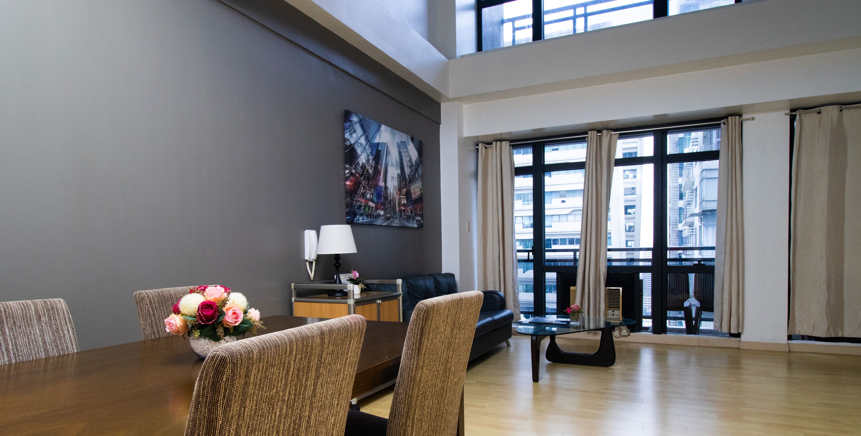 A modern hotel room with a dining area, a sofa, a painting on the wall, and large windows with curtains.