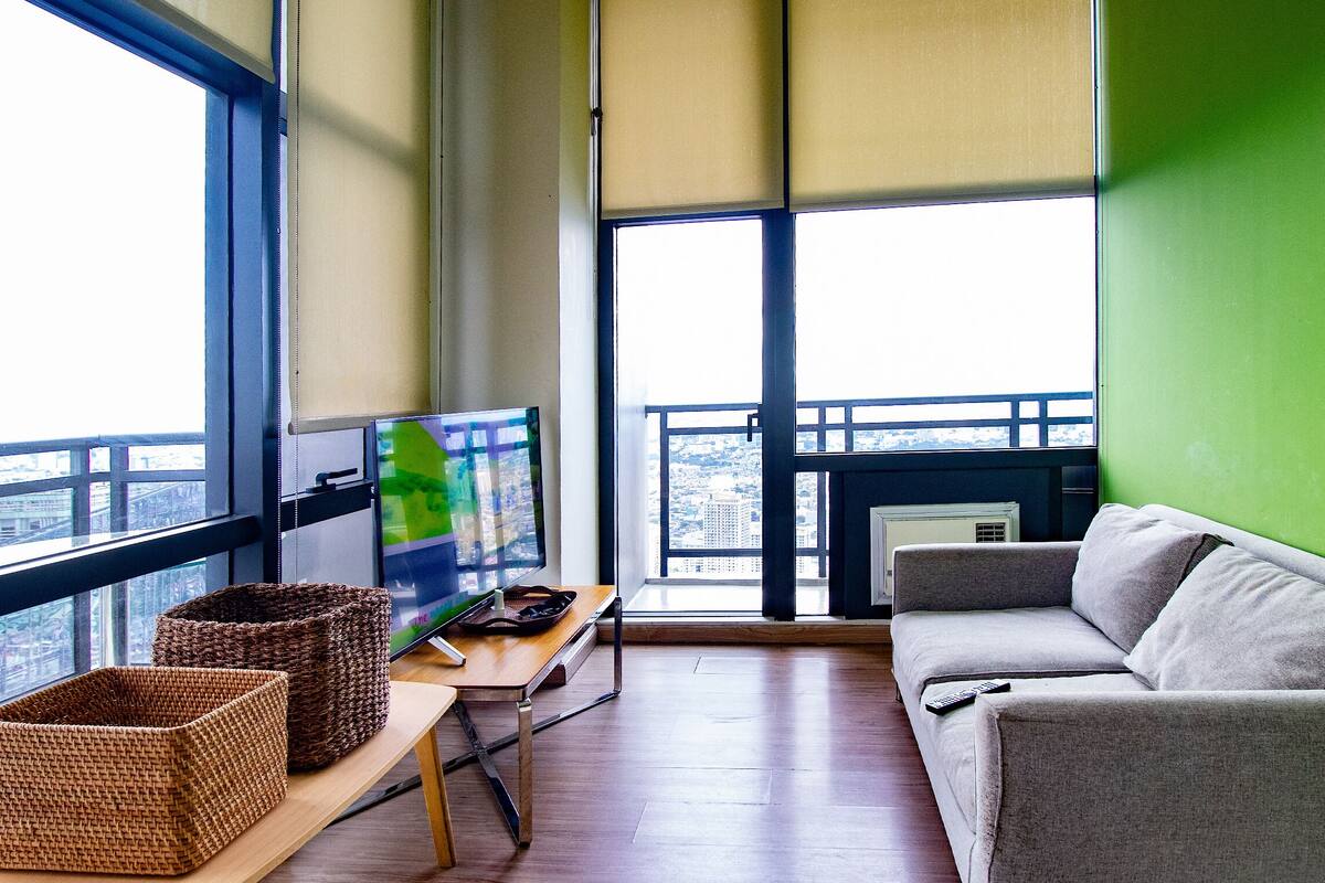 A balcony with a view of a cityscape, a grey sofa, a wooden table with a basket, and a television.