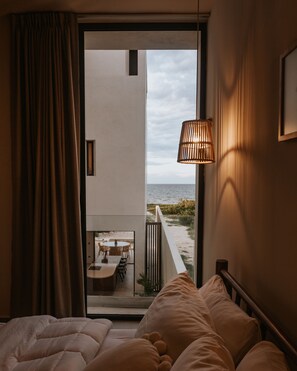 Habitación de hotel con cama, vista a la playa y zona de comedor visible a través de la puerta de vidrio.