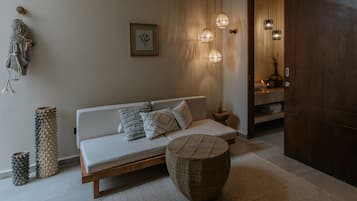 Una sala de estar moderna con un sofá, cojines, un otoman redondo, una alfombra texturizada, un adorno de macramé en la pared y una obra de arte enmarcada.