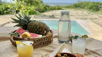 Desayuno frente al mar con frutas tropicales, pan, aceitunas, jamón serrano y un vaso de limonada.