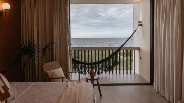 Habitació amb llit, cadira, taula petita amb un ram i un hamaca, amb vista al mar des d'un balcó.
