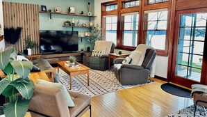 A modern living room with a large flat-screen TV, comfortable seating, and a view of a lake through large windows.