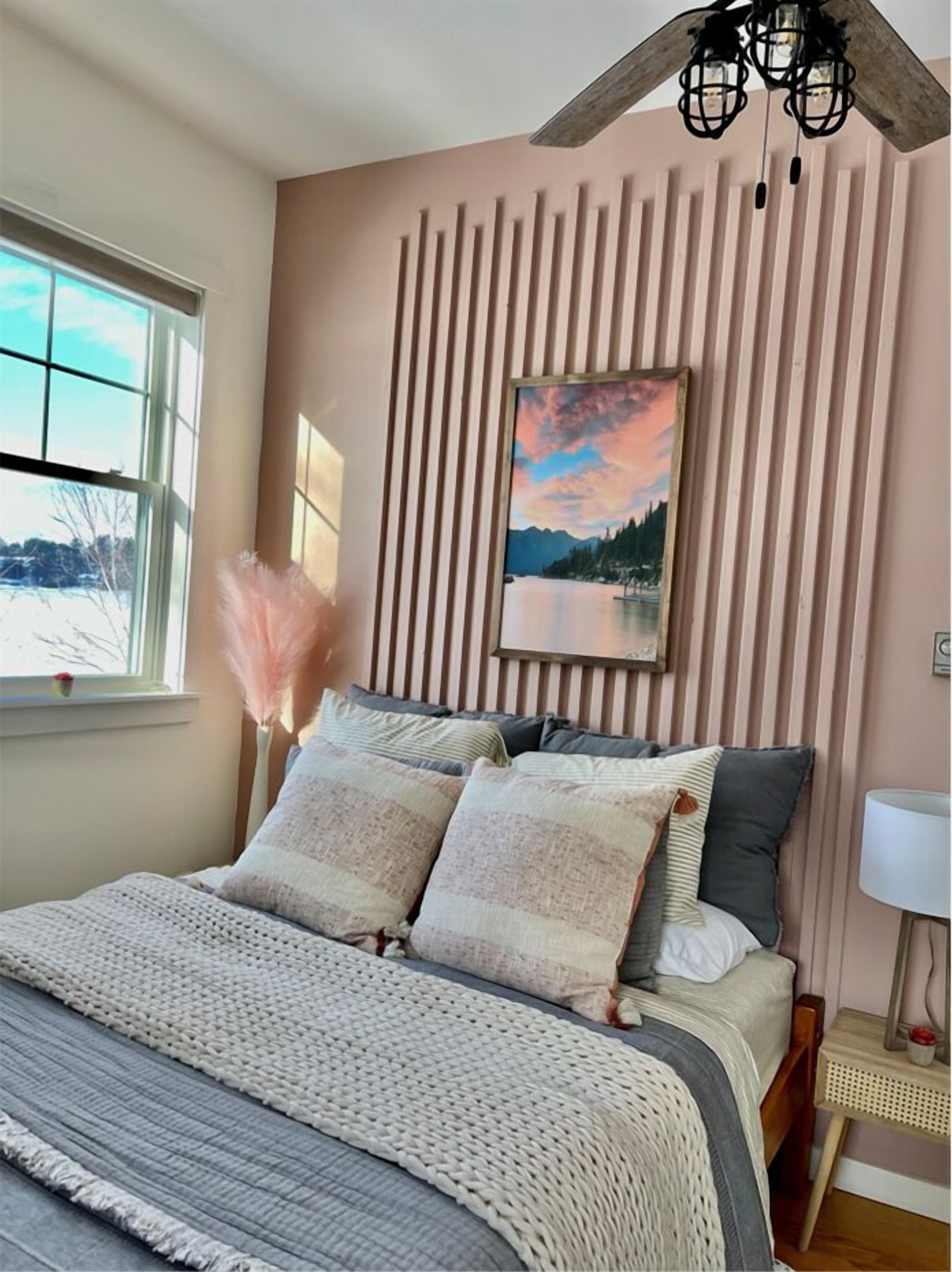A bedroom with a bed, a window, a painting, and a ceiling fan.