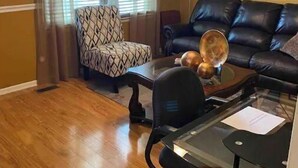 Una sala de estar con un sofá de cuero negro, un sillón estampado, una mesa de centro de vidrio y un ventilador de techo.