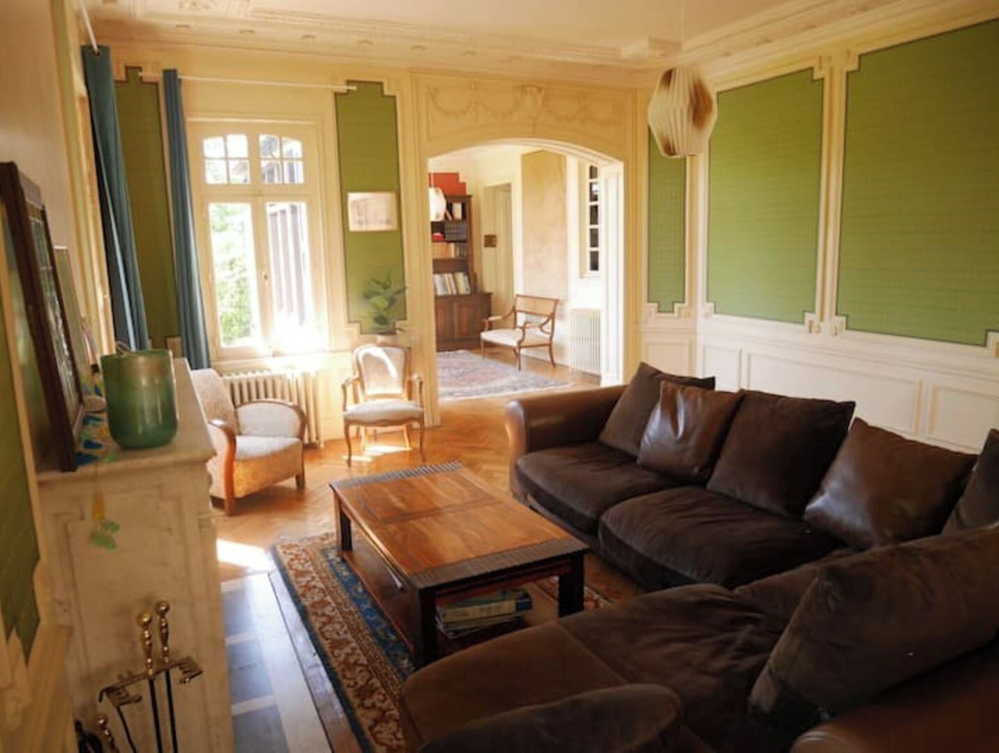 Un salon avec un canapé en cuir marron, une table basse en bois et un fauteuil vert. On aperçoit une bibliothèque et un radiateur au fond.