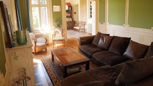 Un salon avec un canapé en cuir marron, une table basse en bois et un fauteuil vert. On aperçoit une bibliothèque et un radiateur au fond.
