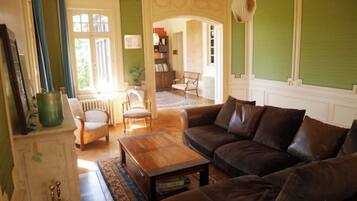 Una sala de estar con un sofá de cuero marrón, una mesa de centro de madera y un sillón verde. Se ve una estantería y un radiador al fondo.