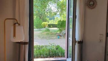 Una habitación con piso de madera, una cama con cabecera, una ventana con cortinas y vista a un jardín.