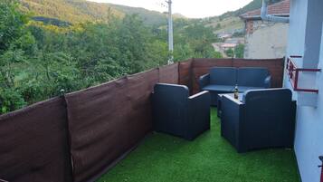 Une terrasse extérieure avec des meubles en osier et une bouteille de bière, entourée d’une clôture marron et d’un gazon synthétique vert.