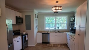 A modern kitchen with white cabinets, stainless steel appliances, a large window, and a hanging light fixture.