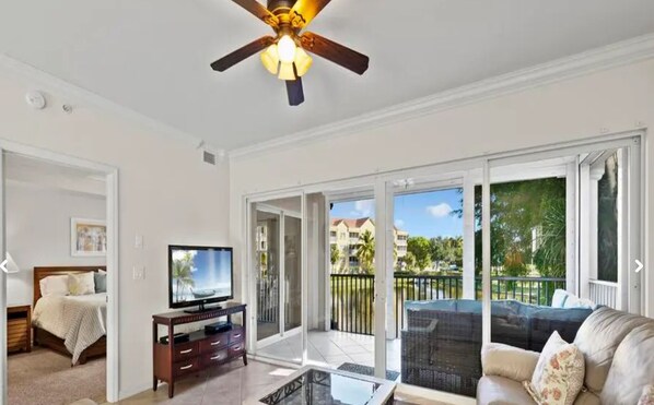 A living room with a TV, a ceiling fan, a sofa, and a balcony with a view of palm trees and buildings.