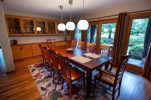 A dining area with a wooden table, chairs, and a floral rug. There are wooden cabinets above the table and a window with curtains.