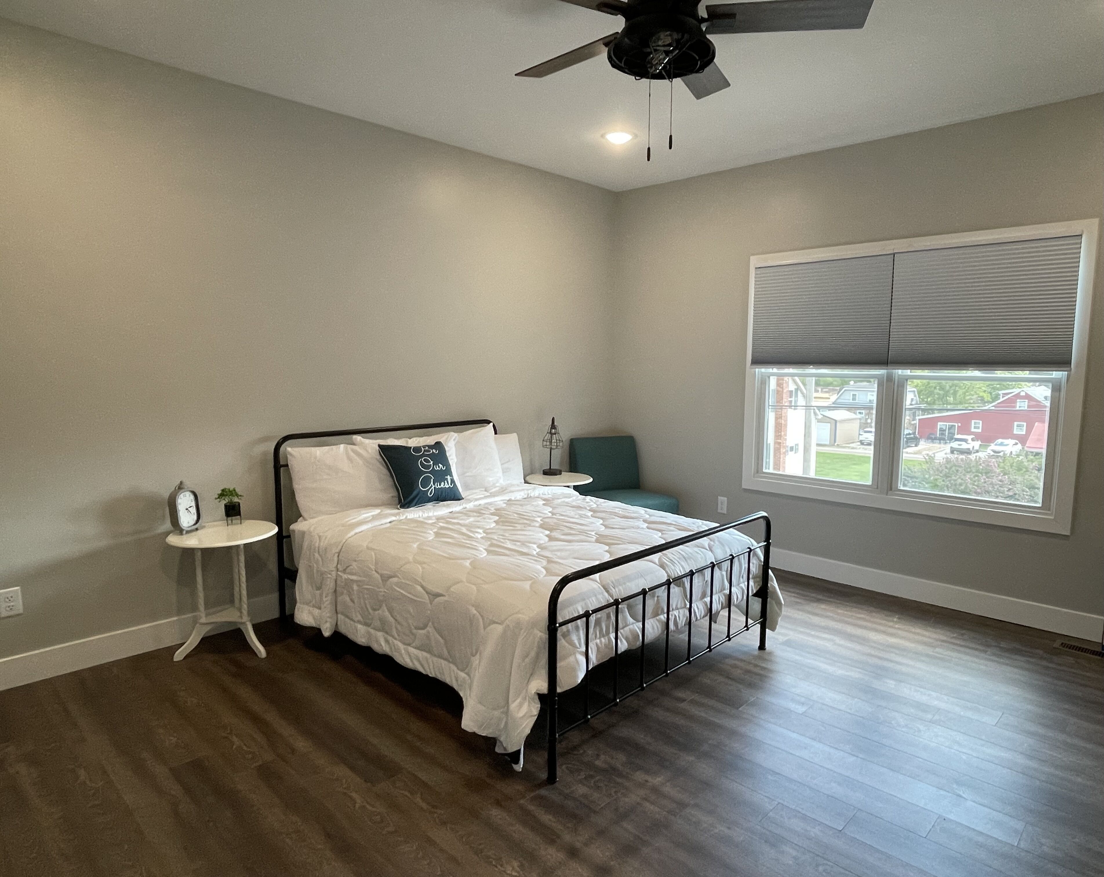 A bedroom with a bed, a nightstand, a small table, a ceiling fan, and a window with blinds.