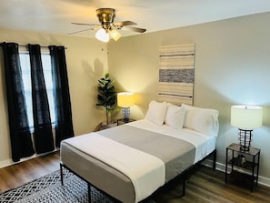 Une chambre d’hôtel comprenant un lit, deux tables de chevet avec des lampes, un ventilateur de plafond, une fenêtre avec des rideaux et une œuvre d’art accrochée au mur.