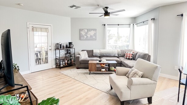 A spacious living room with a sofa, coffee table, and a TV. There is a ceiling fan, a world map on the wall, and a window with curtains.