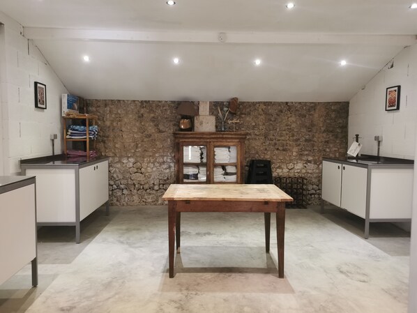 A room with a central wooden table, stone wall, and a kitchenette area with cabinets and a sink. - Chez Foucherie | Luxury Family Countryside Retreat (Nonac)