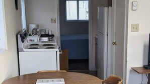 A compact kitchen with a stove, coffee maker, and refrigerator. There is a dining table with two chairs and a book on it. A window with blinds is visible in the background.
