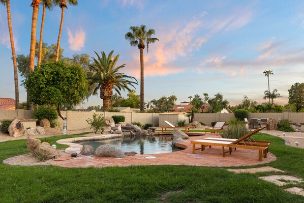 A backyard with a swimming pool, palm trees, and a patio area with lounge chairs.