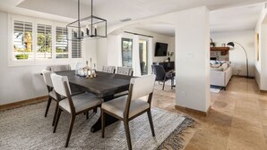 A dining area with a dark wooden table and light-colored chairs, a hanging light fixture, and a view of a garden through large windows. - Sparkling Pool, Poker Table, and Relaxation at Sweetwater (Scottsdale)