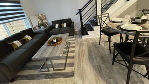 A modern living room with a sofa, coffee table, and dining area with chairs and a table set for a meal. - Minutes to Lake Travis NEW Modern Townhome (Lago Vista)