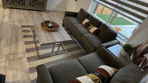 A modern living room with a flat-screen TV, a sectional sofa, a coffee table, and a large window with blinds. - Minutes to Lake Travis NEW Modern Townhome (Lago Vista)