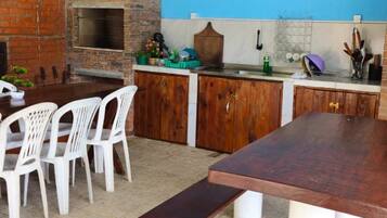 Una cocina al aire libre con horno de ladrillo, sillas de plástico blanco y una mesa de madera.