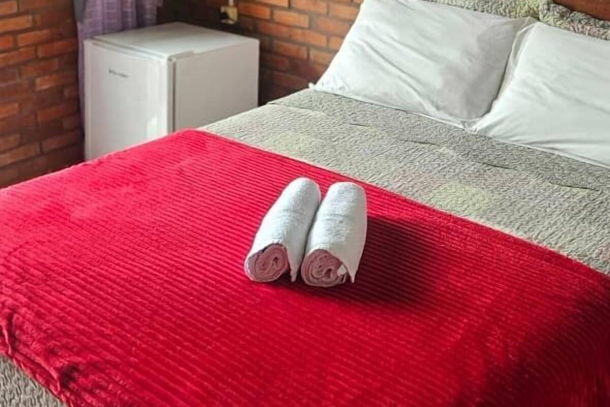 A hotel room with a brick wall, a bed with white and grey bedding, a red blanket, and two rolled towels on the bed.