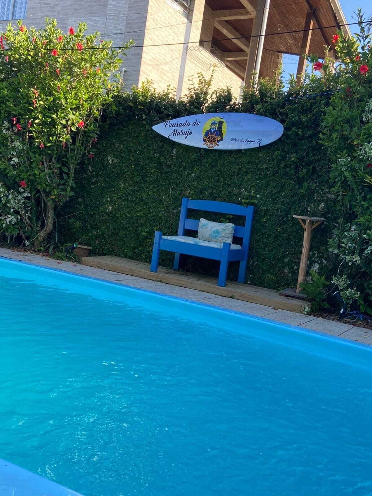 A poolside area with a blue bench, a sign reading "Pensão da Marujo," and a building in the background.