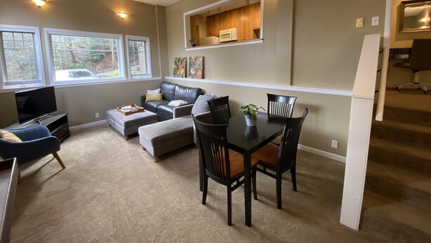 Un salon moderne avec un canapé, un pouf et une table à manger avec des chaises. On peut voir une télévision, une fenêtre et un coin cuisine.