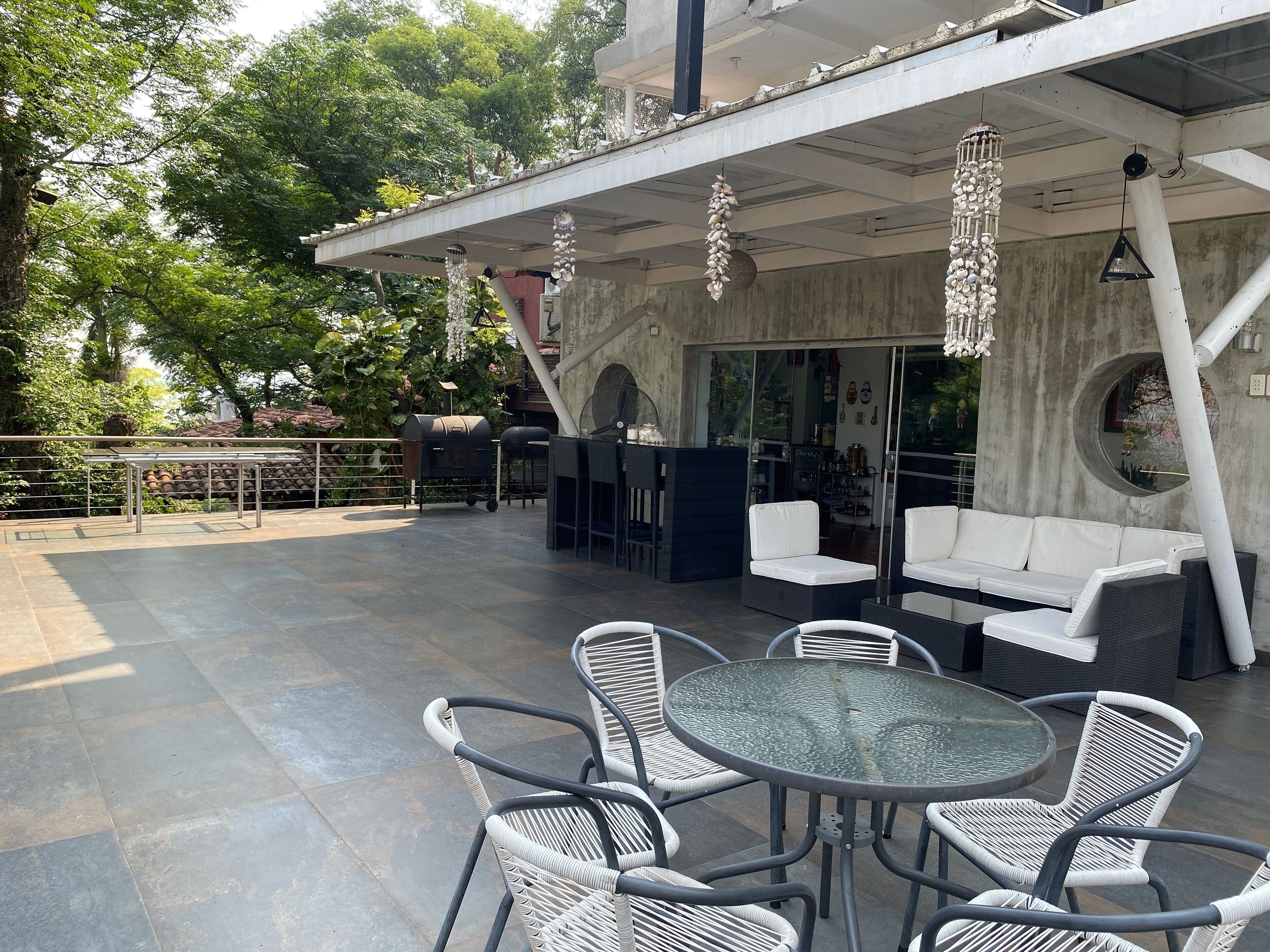 Een terras buiten met een glazen tafel, omringd door rieten stoelen, een witte bank en een barbecue.