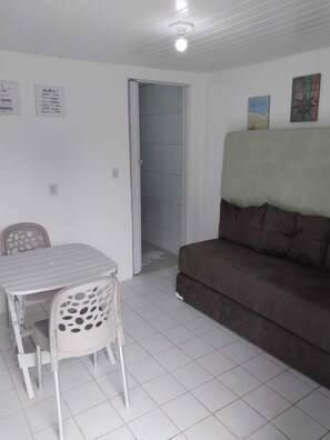 Um quarto pequeno e limpo, com uma mesa e cadeiras brancas, um sofá marrom e uma porta que dá para uma área com azulejos.