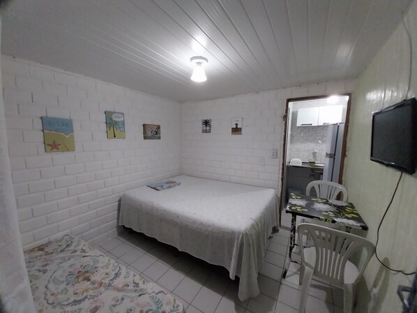 Quarto pequeno com paredes brancas, uma cama de solteiro, uma mesa pequena com cadeira e televisão na parede.