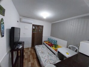 Quarto pequeno e compacto, com cama, escrivaninha, cadeira, televisão e janela com cortinas.